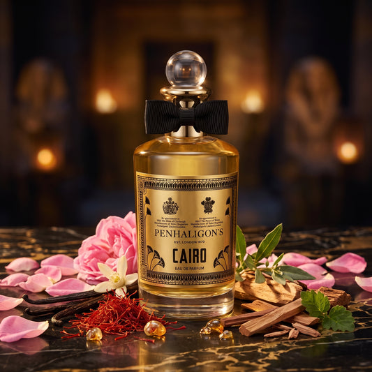 Frasco clásico cilíndrico con líquido dorado, lazo negro y etiqueta dorada con motivos egipcios de Cairo de Penhaligon's, rodeado de rosa de Damasco, azafrán, vainilla y sándalo, estilo Casa Flores.