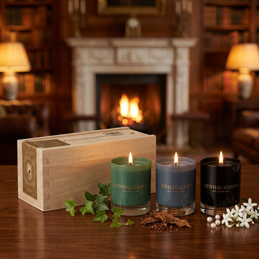 Tres velas encendidas de Penhaligon's en vasos verde, gris y negro con letras doradas, junto a un estuche de madera clara, estilo Casa Flores.