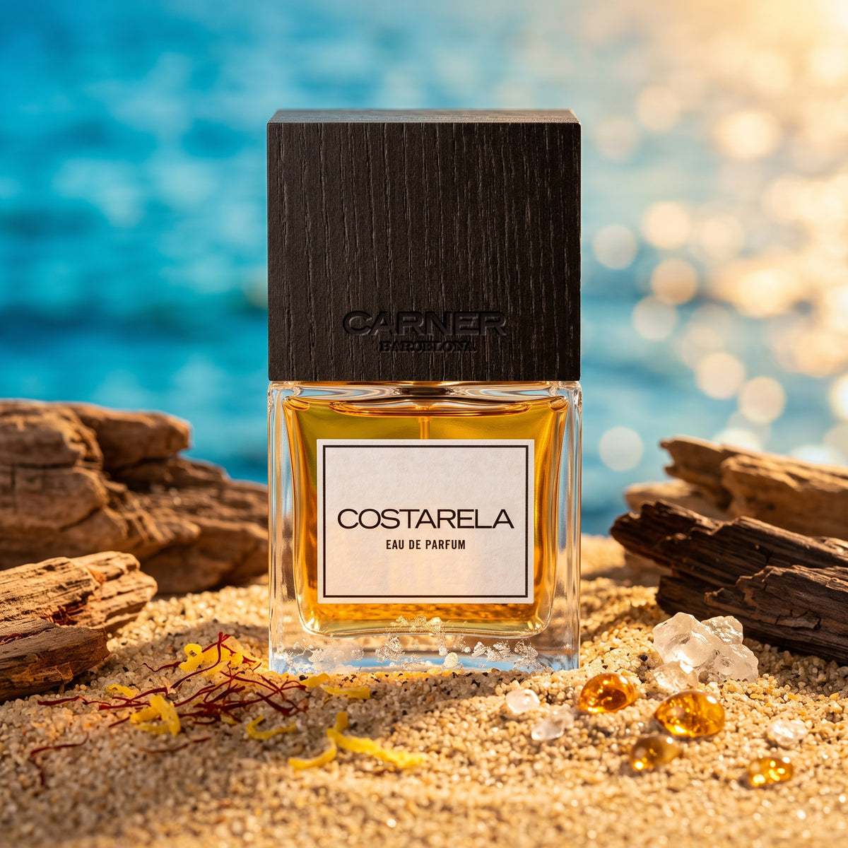 Frasco de cristal con líquido ámbar dorado claro, etiqueta blanca con borde negro y tapón de madera oscura de Costarela de Carner Barcelona, rodeado de arena, notas marinas, bergamota y madera de cedro, estilo Casa Flores.