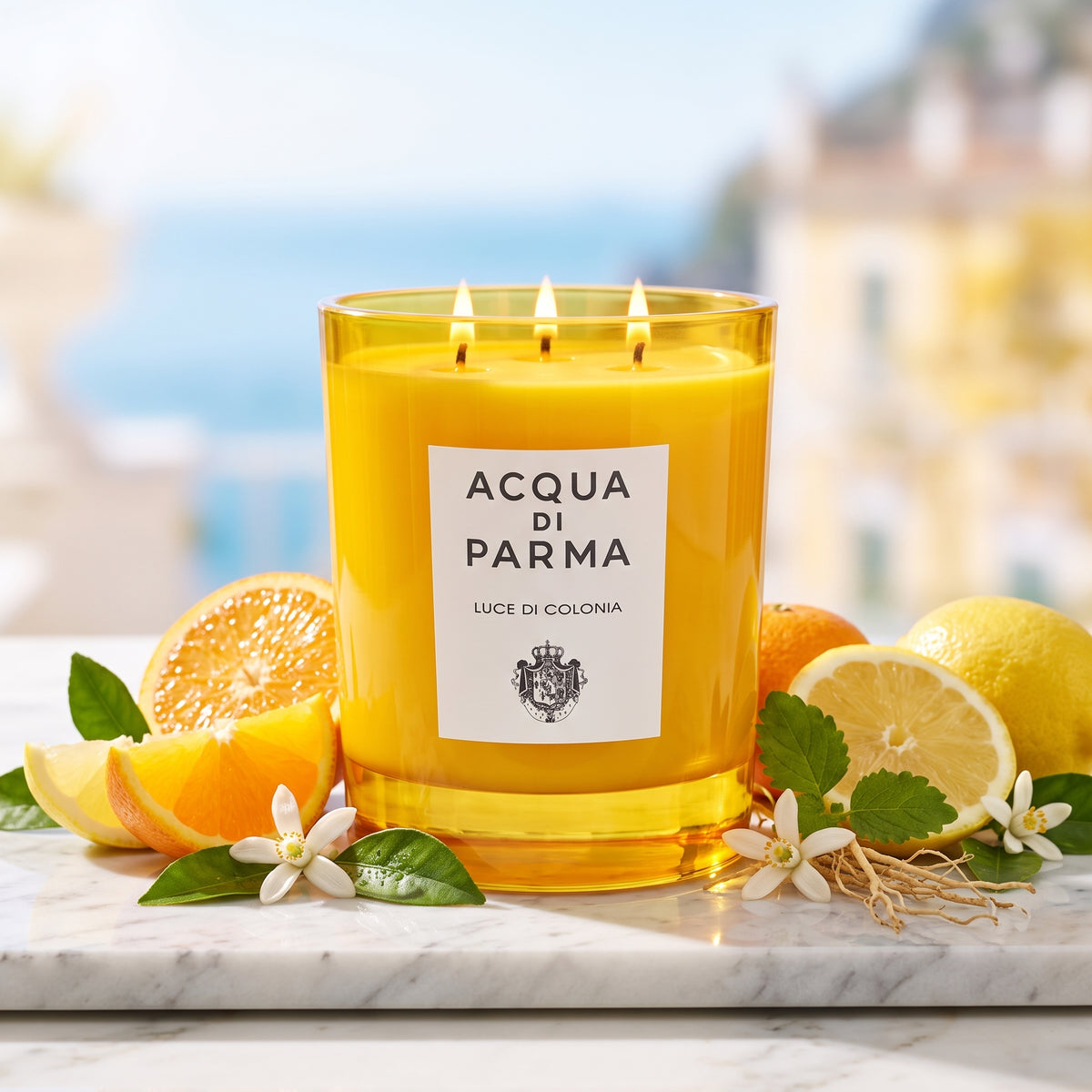 Vela amarilla de cristal con 3 mechas encendidas y etiqueta blanca de Luce di Colonia de Acqua di Parma, rodeada de naranjas, limones y neroli, estilo Casa Flores.