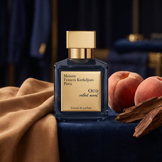 Frasco cuadrado azul muy oscuro con placa dorada y tapón dorado de Oud Velvet Mood de Maison Francis Kurkdjian, rodeado de gamuza, madera de oud y durazno, estilo Casa Flores.