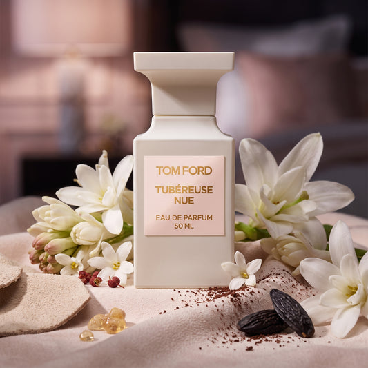 Frasco arquitectónico blanco marfil mate con etiqueta rosa pálido y letras doradas de Tubéreuse Nue de Tom Ford, rodeado de nardos, jazmín, gamuza y cacao, estilo Casa Flores.